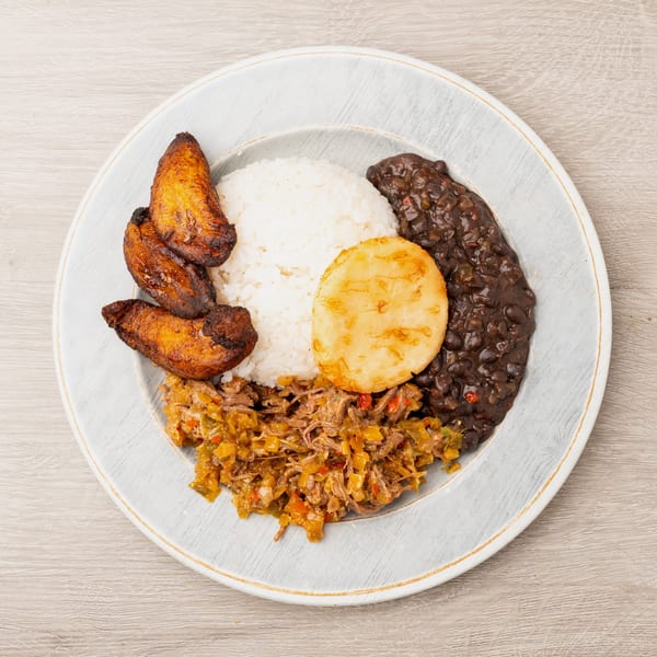A plate of Cuban ropa vieja with rice, black beans, fried plantains, and a corn cake.
