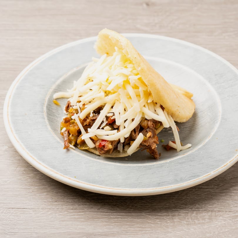 Arepa filled with shredded beef and vegetables, topped with a generous amount of shredded white cheese.