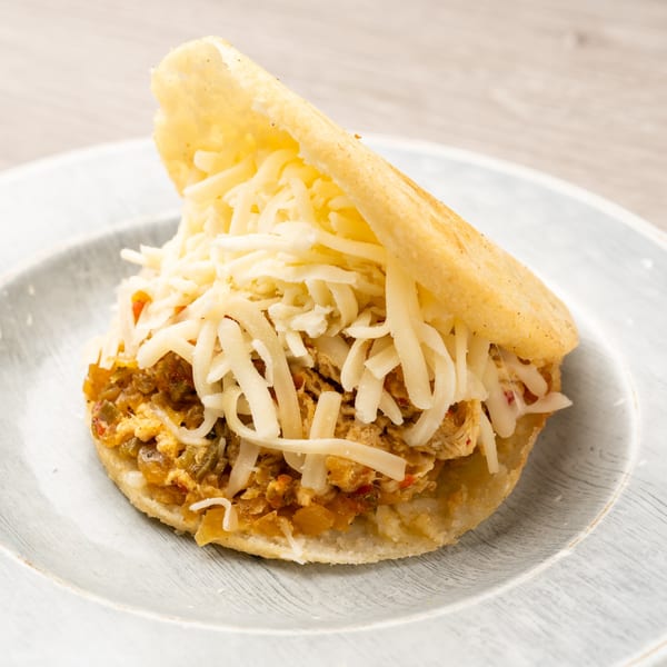 An open arepa filled with shredded chicken and vegetables, topped with shredded white cheese on a plate.